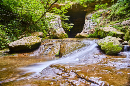 Cave opening inside a forest with green trees and a white flowing river with large bouldersの写真素材