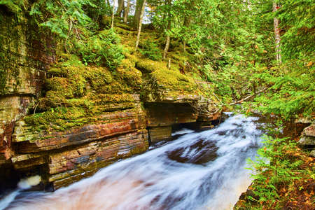 Flowing river of white with cliff wall of stone that looks stacked with moss trees and bushesの写真素材