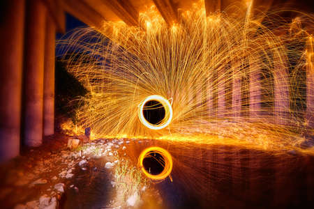Portal of yellow and white sparks of light from steel wool reflected in water under a concrete bridgeの写真素材