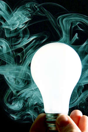 White lightbulb held by a human hand against a background of teal smoke and blackの写真素材