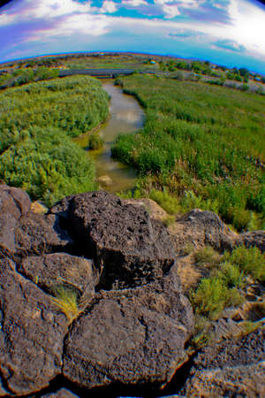 Large black rocks obscure the mouth of a river that flows through green pastures in a fish-eye shotの写真素材