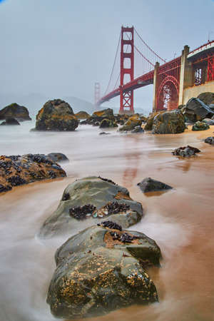 Golden Gate Bridge on foggy morning with smooth waves over tide pools and rocks on beachの写真素材