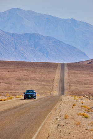 Car driving on long and bare road in empty desert landscape leading to mountainsの写真素材