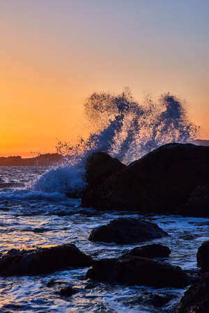 Golden hour on ocean with large wave crashing over rocksの写真素材