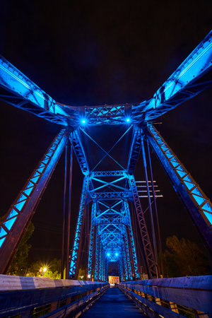 Black sky above glowing neon blue truss railroad bridge with wooden walking bridge