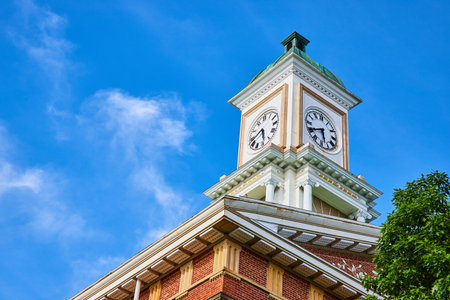 Clock tower on brick building roof with blue skies and green tree topの写真素材