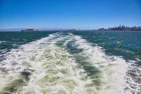 Wake behind boat and choppy water in bay with distant Alcatraz Island and San Francisco skyscrapersの写真素材