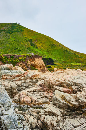 Baren rock cliff face with jagged crags and distant green mountainous hillside under overcast skyの写真素材