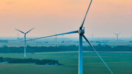 Aerial crisp wind turbine at sunset with orange sky with wind farm behind it and green fields below