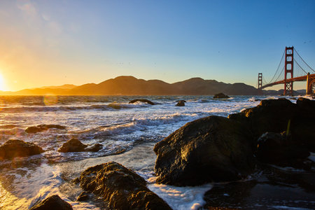 Gorgeous sunset over waves and boulders with Golden Gate Bridge and distant mountainsの写真素材