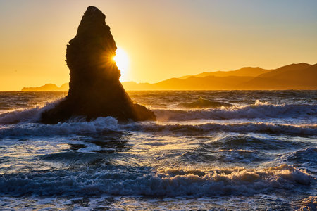 Tall massive boulder jutting out of ocean waves with golden sun poking out from behind