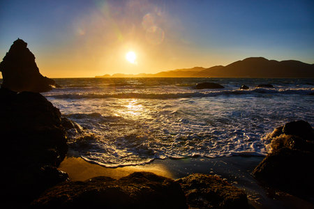Glorious golden sunset over ocean waves rolling into shore with distant mountainsの写真素材