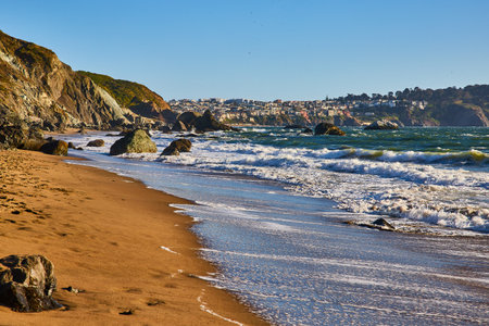 Receding waves on sandy beach with boulders and distant city on top of cliffの写真素材