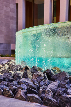 Circular wall with distorted glass from falling water over large black rocks in front of buildingの写真素材