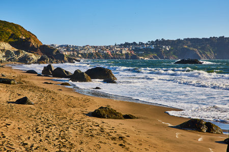 Mossy boulders on beach with tons of footprints in sand and distant city above cliff and wavesの写真素材