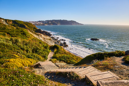 Wooden stairs going down green hill toward rocky shore with small beach and wavesの写真素材