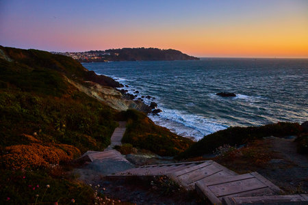Steps going down hill toward blue ocean with sunset sky and distant city above cliffの写真素材