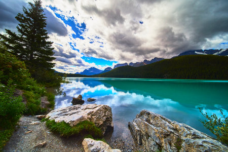 Turquoise Lake with Rocky Shore and Pine Trees against Mountain Backdropの写真素材