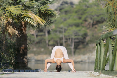 A beautiful woman in a white gymnastic jumpsuit is engaged in yoga, on the beach, against the backdrop of water and a green mountain slopeの写真素材