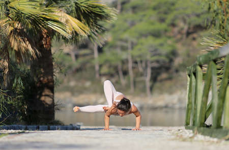 A beautiful woman in a white gymnastic jumpsuit is engaged in yoga, on the beach, against the backdrop of water and a green mountain slopeの写真素材
