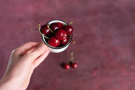 Cup with berries in a female hand. Cherry background. Summer fruits and food concept. With copy space.の写真素材