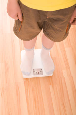 Boy measures weight on floor scales. Legs in shorts and socks standing at floor scales on hardwood floor in living room. View from above.の写真素材