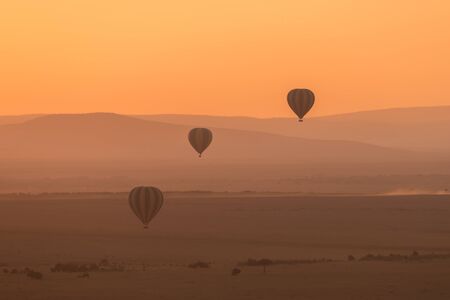 Three hot air balloons fly over the low hills of the African savannah. The aerial perspective makes the hills look different shades of purple, and the sky is bright orange in the pre-dawn light.の写真素材