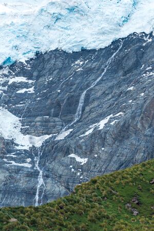Waterfall on cliff between glacier and treesの写真素材