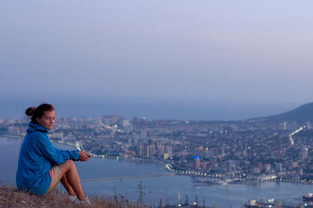 a girl in a blue sweater sits on a mountain overlooking the city, the concept of hiking in the...