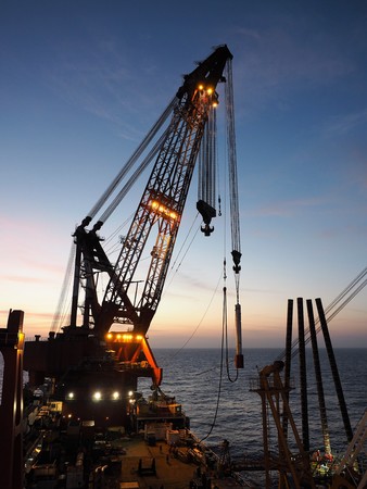 Derrick barge's crane lifts piling hammer to drive piles during platform jacket installation.