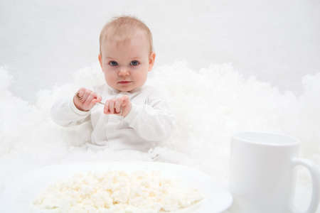 cute little girl sitting on white blanket with spoon in her hands. There are white plate with curd and white cup in front of her. Soft-focused, focal point is on the girlの写真素材