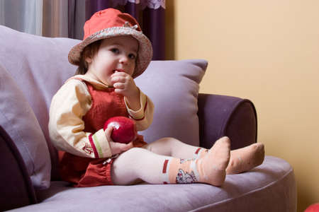 cute little girl with red apple in her hand sitting on the sofaの写真素材