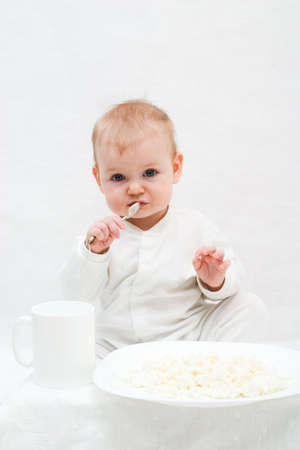cute little girl sitting on white blanket with spoon in her hands. There are white plate with curd and white cup in front of her.の写真素材