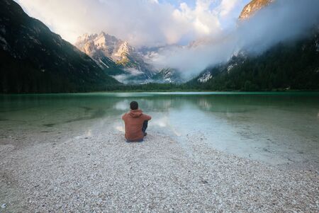 Man sitting near mountain lake at cloudy morning. Lago di Landro, Dolomites Alps, Italyの写真素材