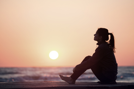 Lonely girl sitting on the sunrise beach. Woman silhouette over sunrise skyの写真素材