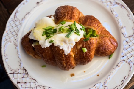 Delicious croissant with bacon and scrambled egg served on a plate on a wooden table.