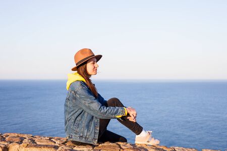 Closeup back view of woman in jeans jacket and hat sitting and looking at blue ocean and sky.の写真素材