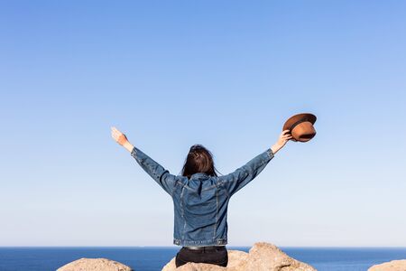 Closeup back view of woman in jeans jacket and hat sitting and looking at blue ocean and sky.の写真素材