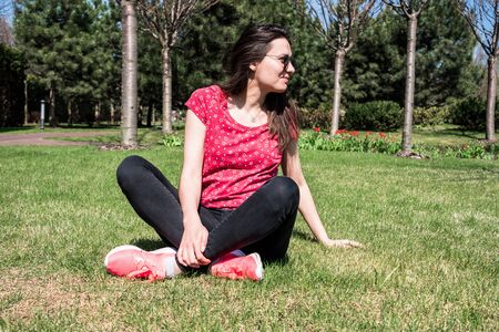 Cute long-haired girl sitting on the green lawn and looks to the side. Girl in the city spring parkの写真素材