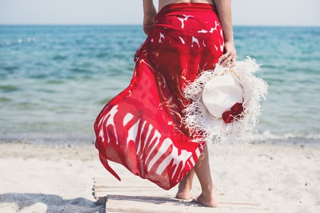 Beautiful woman with white hat walks at the sea side in red dressの写真素材