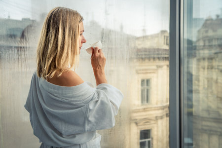 blonde woman in bathrobe using tissue while standing near window at homeの写真素材