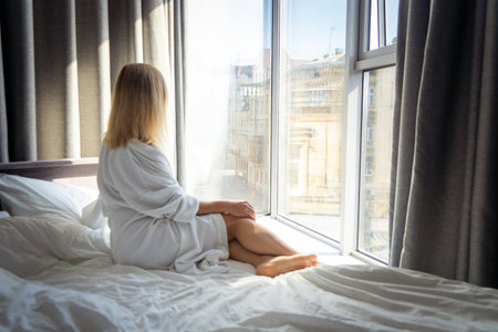 young woman in bathrobe sitting on bed and looking out the windowの写真素材