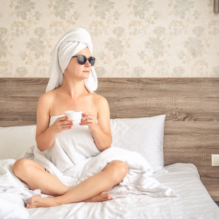Young beautiful woman with a towel on her head sitting on the bed with a cup of coffee.の写真素材