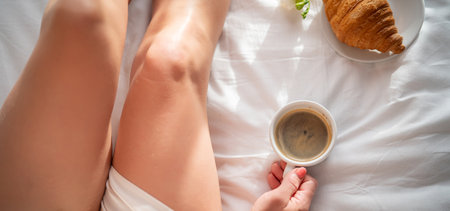 cropped shot of woman holding cup of coffee and croissant on bedの写真素材
