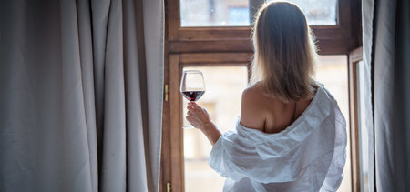 back view of woman holding glass of red wine in bedroom at homeの写真素材