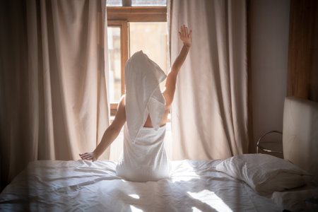 Back view of young woman in towel sitting on bed and meditatingの写真素材