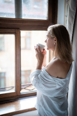 Woman in elegant robe drinking coffee and standing near windowの写真素材