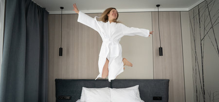 Woman having fun and jumping with bathrobe on bed in hotel roomの写真素材