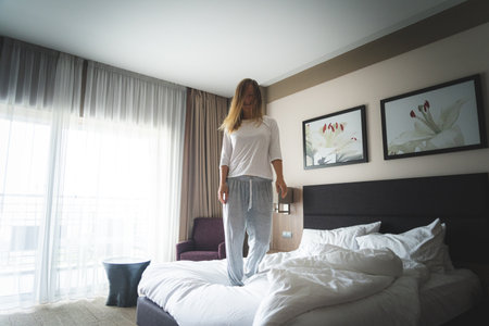 beautiful female dropping pilows and jumping with bathrobe on bed in hotel room