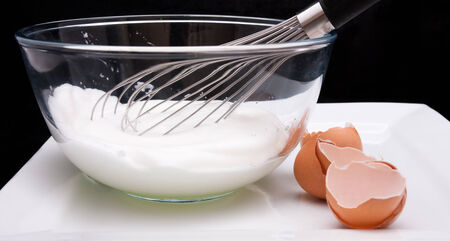 Egg whisk in a bowl with Egg whites and broken egg shells against a black backgroundの写真素材